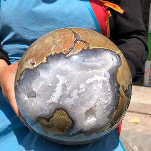May include: A polished spherical stone featuring a large, central geode cavity filled with white crystalline structures. The outer surface displays swirling patterns of brown, tan, and dark gray mineral bands surrounding the central white quartz.