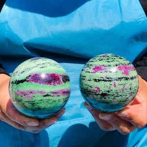 May include: Two polished spherical stones featuring a vibrant mix of light green, deep magenta, and black mineral inclusions. The spheres have a smooth, reflective surface with distinct horizontal bands of color throughout their rounded forms.