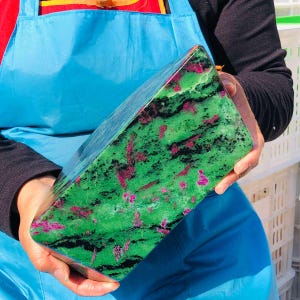 May include: A large, rectangular block of polished zoisite mineral featuring a vibrant green base color with prominent inclusions of deep magenta ruby crystals and dark black hornblende streaks throughout the stone's smooth, reflective surface.