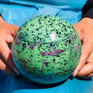 May include: A polished spherical stone featuring a vibrant green base color with scattered black speckles and distinct patches of deep magenta-pink mineral inclusions throughout the smooth, rounded surface of the orb-shaped mineral specimen.