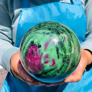 May include: A polished spherical stone featuring a vibrant mix of bright green and deep magenta-pink mineral inclusions, accented by dark black streaks and swirls throughout the smooth, rounded surface of the orb-shaped mineral specimen.