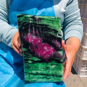 May include: A large, polished slab of zoisite mineral featuring a prominent central patch of vibrant magenta ruby surrounded by deep green zoisite and streaks of black hornblende. The stone has a smooth, rectangular shape with rounded edges.