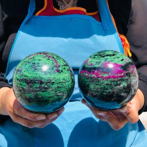 May include: Two polished spherical stones featuring a vibrant mix of green zoisite, deep black hornblende, and bright magenta ruby inclusions. Each sphere displays a smooth, reflective surface with natural color variations and distinct mineral patterns.