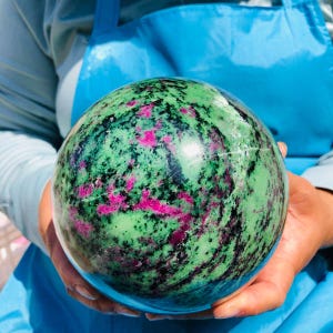 May include: A polished spherical stone featuring a vibrant mix of mottled green, deep black, and bright magenta-pink mineral inclusions. The surface is smooth and reflective, showcasing a natural marbled pattern throughout the entire round object.