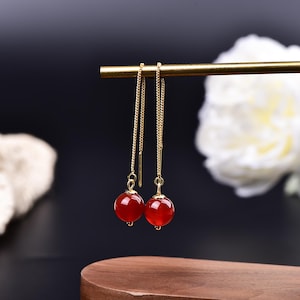 May include: A pair of gold-colored chain earrings with red, round gemstone beads. The earrings hang from a gold bar, with a dark background and a white flower in the background. The earrings are displayed on a wooden surface.