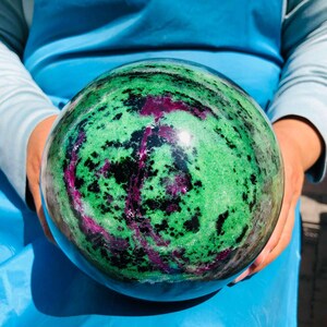 May include: A polished spherical stone featuring a vibrant mix of bright green, deep black, and magenta-colored mineral inclusions. The surface is smooth and reflective, displaying a swirling, organic pattern of contrasting colors throughout.