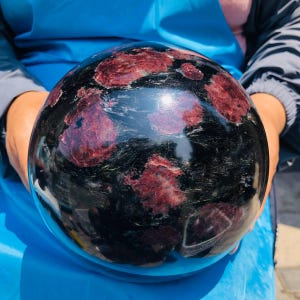 May include: A polished spherical mineral specimen featuring a deep black base color with prominent, irregular inclusions of dark red garnet crystals throughout the surface, showcasing a smooth, reflective, and glossy finish across the entire sphere.