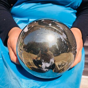 May include: A polished, spherical pyrite stone with a highly reflective, metallic gold surface. The sphere features natural, irregular white mineral inclusions and a smooth, mirror-like finish that reflects the surrounding outdoor environment.