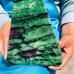 May include: A triangular slab of zoisite mineral featuring a vibrant green base color with prominent black hornblende inclusions and scattered deep magenta ruby patches throughout the polished stone surface, displaying natural crystalline patterns.