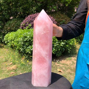 May include: A large, polished rose quartz crystal tower with a hexagonal base and a pointed top. The stone features a translucent, pale pink color with natural internal veining and cloudy white inclusions throughout its solid, vertical structure.