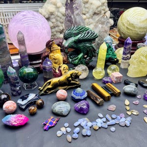 Puede incluir: Una colección de varios cristales y piedras preciosas, que incluye esferas, obeliscos y figuras talladas. El surtido presenta una gran esfera de color púrpura claro, un dragón verde tallado y una calavera amarilla. Otras piedras incluyen ojo de tigre, amatista y fluorita.