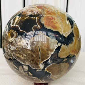 May include: A polished spherical stone featuring a complex pattern of deep black, earthy brown, cream, and reddish-orange patches. The surface is highly reflective, showing light reflections and intricate mineral inclusions throughout the orb.
