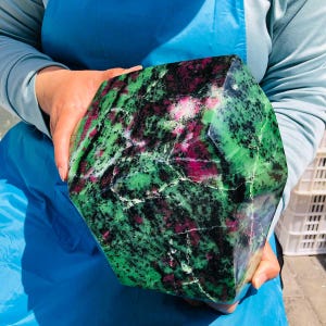 May include: A large, polished, multi-faceted mineral specimen featuring a vibrant mix of deep magenta-pink ruby inclusions embedded within a matrix of bright green zoisite and scattered black hornblende mineral specks throughout the stone.