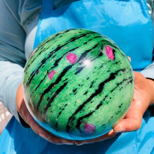 May include: A polished spherical stone featuring a vibrant green base color with prominent black streaks running across the surface and scattered, irregular bright pink inclusions throughout the entire round mineral specimen.