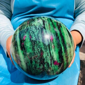 May include: A polished spherical stone featuring a vibrant green base with prominent black streaks and scattered magenta-colored inclusions throughout the surface. The sphere displays a smooth, reflective texture with natural mineral patterns.