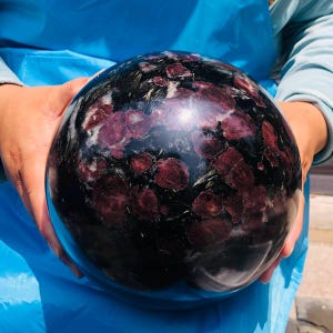 May include: A polished spherical stone featuring a deep black base color with prominent dark red, circular inclusions and thin, needle-like metallic streaks scattered throughout the surface, creating a complex, multi-textured appearance.