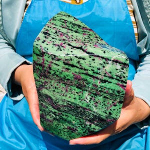 May include: A large, polished stone with a vibrant green base color, speckled with black and magenta inclusions. The stone is held in two hands, showcasing its irregular, geometric shape. The overall appearance suggests a natural, decorative item.