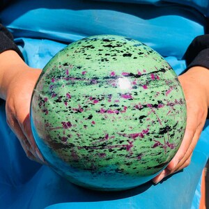 May include: A polished spherical stone featuring a vibrant light green base color with scattered dark black mineral inclusions and distinct magenta-colored crystalline patches distributed across the smooth, rounded surface of the orb.