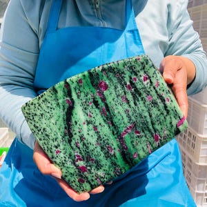 May include: A rectangular slab of polished zoisite stone featuring a vibrant green base with prominent black hornblende streaks and scattered, deep magenta-colored ruby inclusions throughout the smooth, flat surface of the mineral specimen.
