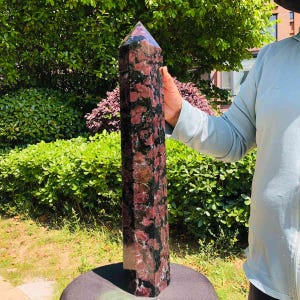 May include: A tall, six-sided crystal tower featuring a mottled pattern of deep black and vibrant reddish-pink mineral inclusions. The stone has a polished, smooth surface with a pointed, faceted top, displaying natural variations in color throughout.
