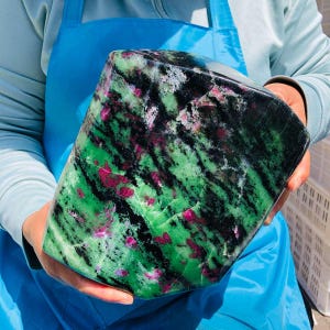 May include: A large, polished slab of zoisite mineral featuring a vibrant mix of deep green, black, and bright magenta-pink inclusions. The stone has a smooth, rounded rectangular shape with visible natural crystalline patterns throughout its surface.