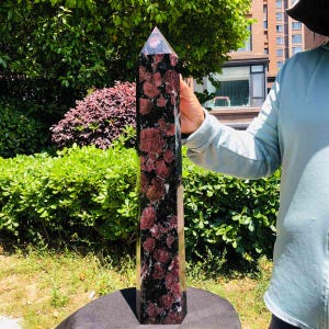 May include: A tall, polished crystal tower featuring a dark black base with prominent, irregular deep red and maroon inclusions throughout its vertical structure, terminating in a sharp, multi-faceted point at the top of the stone column.