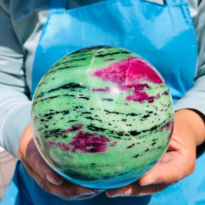 May include: A polished spherical stone featuring a vibrant green base color with prominent patches of deep magenta and streaks of black mineral inclusions throughout the surface, showcasing a smooth, rounded texture and natural color variations.