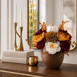 Op de afbeelding: Een bloemstuk in een bruine kom met een witte pompoen, oranje bessen en bloemen in wit, geel en donkerrood. Daarnaast staan twee witte kaarsen in een gouden houder en twee boeken met de tekst CRISPART CATALOGUE op de kaft.