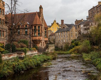 May include: A scenic view of a river flowing through a historic European town. Buildings with stone facades and red-tiled roofs line the riverbanks. Lush greenery and trees add color to the overcast day.