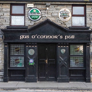 May include: A black storefront with a sign that reads "Gus O'Connor's Pub". The storefront has two windows with stained glass and a double door entrance. There are two beer signs above the entrance, one for Heineken and one for Murphy's Irish Stout.