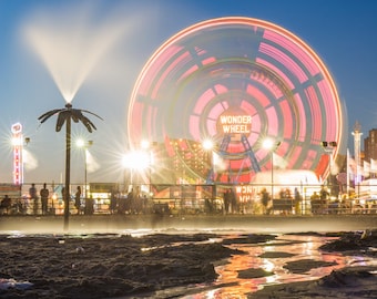 May include: A brightly lit Ferris wheel with the words "Wonder Wheel" on it, seen from a beach at night. The wheel is reflected in puddles of water on the sand.