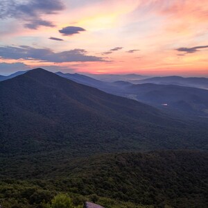 May include: A panoramic view of a mountain range at sunrise. The sky is a vibrant pink and orange, with clouds casting long shadows across the forested slopes.