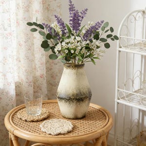 May include: A decorative vase with a cream and brown ombre design holds a bouquet of lavender, white flowers, and green leaves. The vase sits on a round, woven table with two crochet coasters and a clear glass.