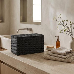May include: A rectangular black woven storage box with a lid, featuring a textured grid pattern. The box is constructed from dark, tightly braided material and sits as a standalone container for organizing items in a bathroom or living space.