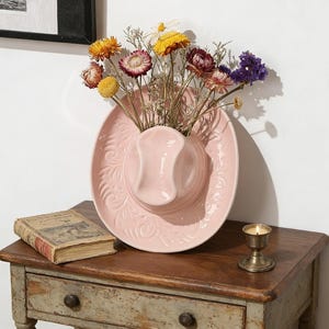 May include: A pink ceramic cowboy hat-shaped wall vase filled with dried flowers in various colors. The vase is mounted on a white wall above a wooden table. A small antique book and a candle in a brass holder are on the table.