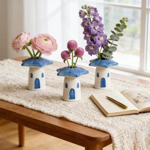 May include: Three white ceramic vases with blue star-shaped tops and arched door designs, each holding different flowers. The vases sit on a cream-colored table runner, with an open book and pen nearby. The flowers include pink ranunculus, purple delphiniums, and pink globe amaranth.