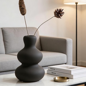 May include: A matte black, gourd-shaped vase with two dried floral stems. The vase sits on a white marble coffee table, with a stack of books and a small gold tray. A gray sofa and a floor lamp are in the background.
