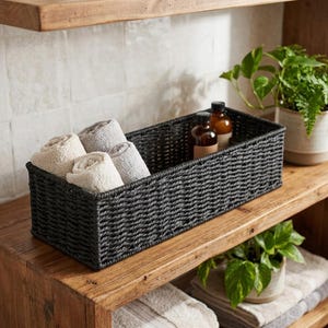 May include: A rectangular black woven storage basket containing four rolled towels in shades of beige and gray. Two amber glass bottles with black caps are positioned inside the basket next to the towels, creating a functional storage set.