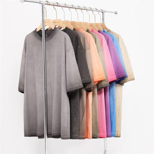 May include: A row of oversized t-shirts in various colors, including gray, black, orange, pink, purple, blue, and beige, hanging on a metal clothing rack. The shirts have short sleeves and a faded, vintage look.