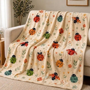 May include: A beige throw blanket draped over a sofa, featuring a repeating pattern of colorful ladybugs, flowers, and green leaves. The ladybugs are red, orange, green, and blue with black spots. The blanket is made of a soft material.