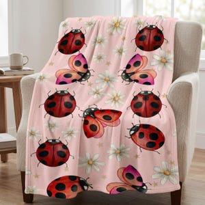 May include: A pink throw blanket featuring a repeating pattern of red ladybugs with black spots, white daisies, and gold stars. The ladybugs have open and closed wings. The blanket is draped over a beige armchair.