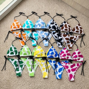 May include: A collection of colorful bikinis with cow print patterns. The triangle tops and string bottoms come in various colors, including orange, teal, blue, black, brown, green, yellow, and pink. Each bikini has black string ties.