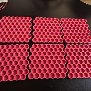 May include: Six square, hot pink silicone mats featuring a repeating honeycomb pattern of circular indentations. Each mat has a grid of small, uniform, recessed circles arranged in a structured, geometric layout across the entire surface.