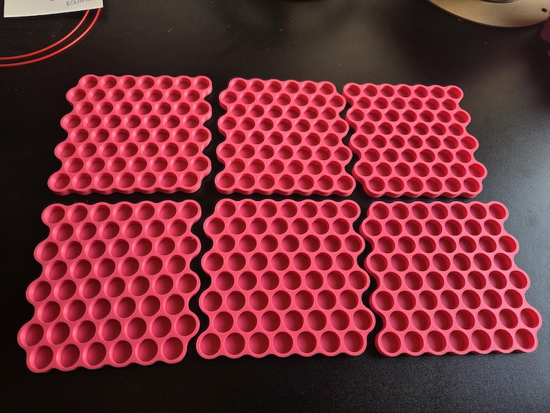 May include: Six square, hot pink silicone mats featuring a repeating honeycomb pattern of circular indentations. Each mat has a grid of small, uniform, recessed circles arranged in a structured, geometric layout across the entire surface.