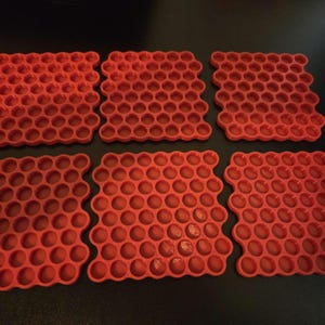 May include: Six square red coasters featuring a repeating honeycomb pattern of circular indentations. Each coaster has a grid of small, uniform, recessed circles arranged in a dense, interlocking hexagonal structure across the entire surface.