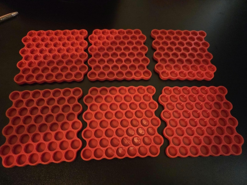 May include: Six square red coasters featuring a repeating honeycomb pattern of circular indentations. Each coaster has a grid of small, uniform, recessed circles arranged in a dense, interlocking hexagonal structure across the entire surface.