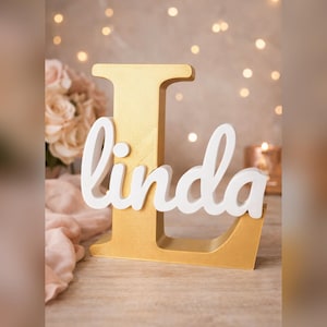 May include: A gold-painted wooden letter "L" with the name "Linda" in white script. The decorative piece is set on a light-colored surface, with soft lighting and blurred background elements including flowers and candles.