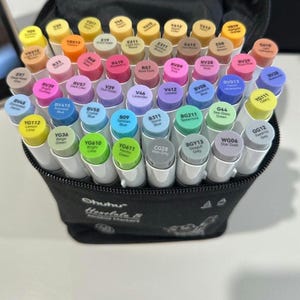 Op de afbeelding: Een zwarte etui met een set Ohuhu alcoholstiften. De stiften hebben witte barrels en gekleurde doppen, met een verscheidenheid aan tinten, waaronder geel, roze, blauw en groen. De etui is open en toont de stiften.