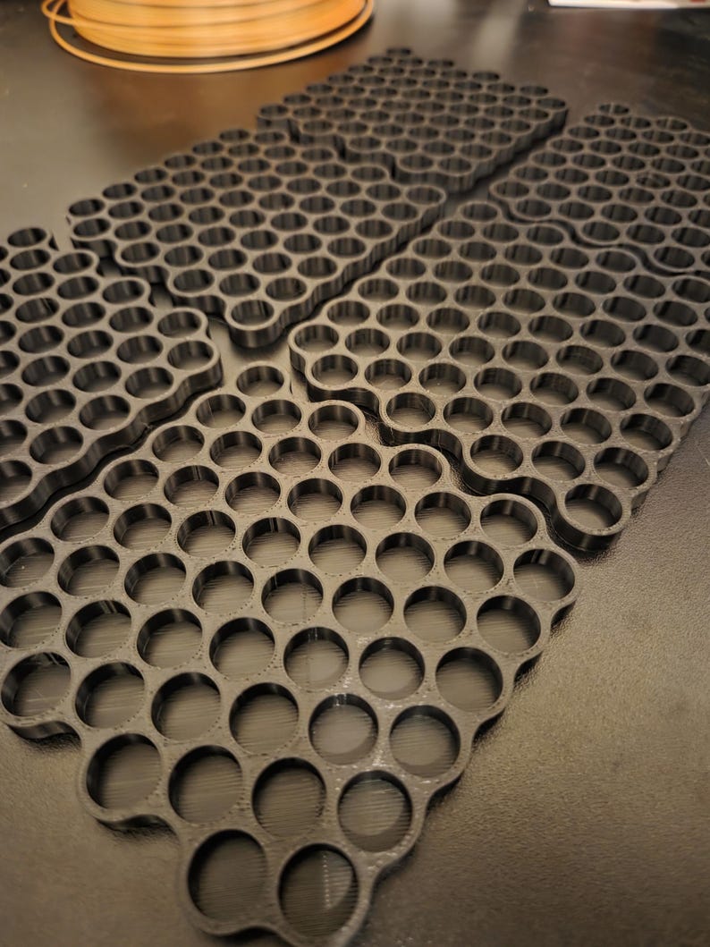 May include: A set of four black 3D printed grid organizers, each featuring a repeating pattern of circular cutouts arranged in a rectangular formation. The organizers are flat with uniform thickness and have a matte finish, designed for storage.