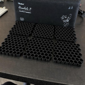 Puede incluir: Una funda negra con la inscripción "Ohuhu Honolulu B Alcohol Markers" y una ilustración de una rana. Debajo, hay seis soportes negros para rotuladores. Cada soporte está diseñado para contener varios rotuladores. La funda tiene capacidad para 320 rotuladores.